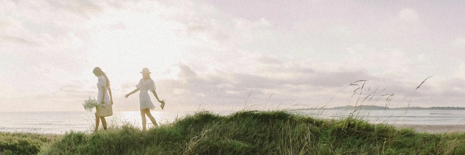Zwei Frauen gehen durch hohes Gras am Strand entlang.